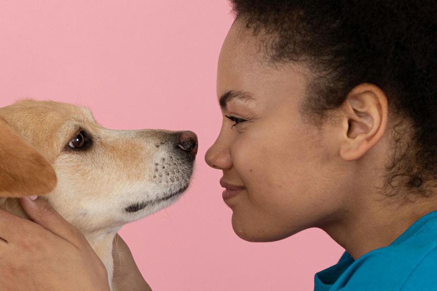 A woman with brown skin and black curly hair smiles nose to nose with a tan dog in front of a pink background.