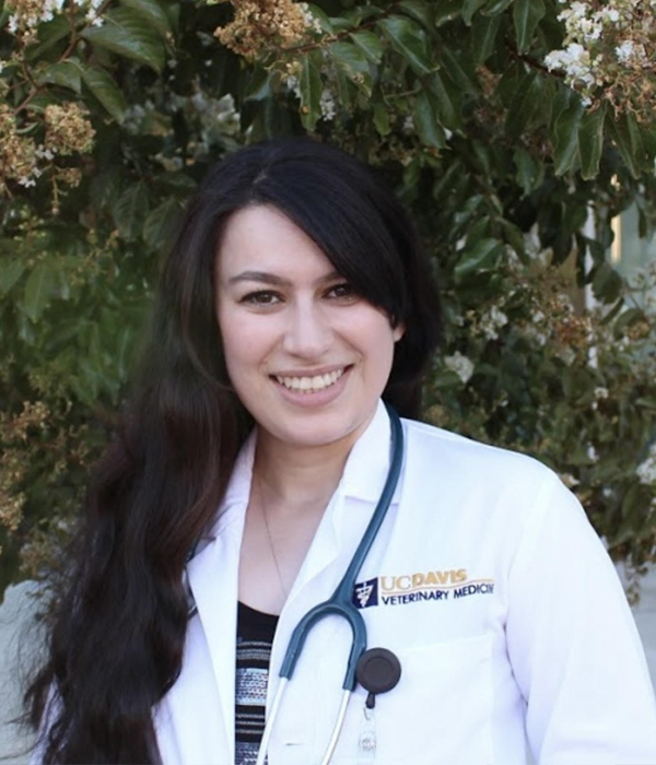 Dr. Haleh Golestani, a female veterinarian with long, brown hair smiles at the viewer from a background of greenery.